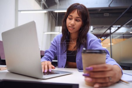 Foto de Mujer concentrada haciendo tareas de trabajo con computadora portátil mientras sostiene la taza de café en la oficina moderna - Imagen libre de derechos