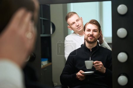 Téléchargez les photos : Joyeux mâle avec une tasse de café en regardant l'auto réflexion dans le miroir tout en coiffeur examiner les cheveux de son client à l'intérieur - en image libre de droit