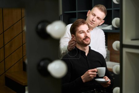 Téléchargez les photos : Réflexion dans le miroir de coiffeur assidu coiffure coupe de cheveux du client qui tient une tasse de café et assis dans le salon de coiffure - en image libre de droit