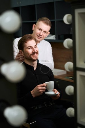 Photo for Young man with coffee cup in his hands smiling while joyful barber styling his haircut indoors - Royalty Free Image