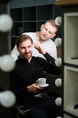 Photo for Confident man holding coffee cup and telling to hairdresser about haircut he wants at barber shop - Royalty Free Image