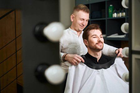 Photo for Reflection in mirror of joyful barber putting on cutting hair cape on his smiling client indoors - Royalty Free Image