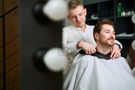 Photo for Good looking client looking at self in mirror while barber preparing him for haircut in modern salon - Royalty Free Image