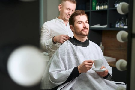 Photo for Confident male client drinking coffee while hairdresser serving him in modern barbershop - Royalty Free Image