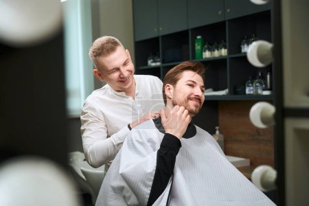 Photo for Mirror reflection of skilled hair stylist laughing while serving male client in white cutting hair cape indoors - Royalty Free Image