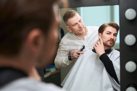 Photo for Male client in cutting hair cape talking to barber about his look while sitting on chair in barbershop - Royalty Free Image