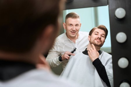 Téléchargez les photos : Coiffeur assidu et beau client se regardant dans le miroir dans le salon de coiffure moderne - en image libre de droit