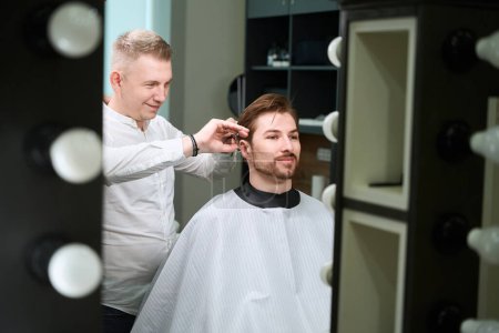 Photo for Smiling hairdresser in shirt cutting hair of his male client by scissors in modern room - Royalty Free Image