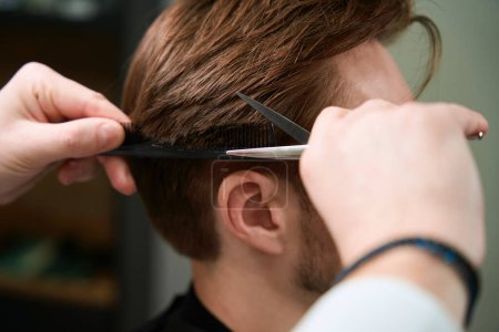 Téléchargez les photos : Vue rapprochée sur les mains du coiffeur couper les cheveux du visiteur à l'aide de ciseaux et peigne à l'intérieur - en image libre de droit