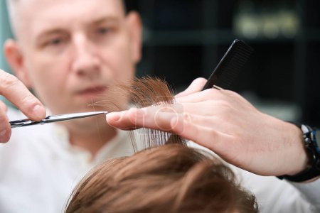 Téléchargez les photos : Coiffeur professionnel travaillant avec des ciseaux et peigne tout en coupant le client dans le salon de coiffure - en image libre de droit