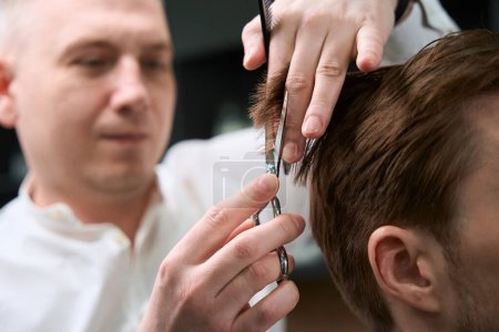 Téléchargez les photos : Coiffeur professionnel en chemise faisant coupe de cheveux à son visiteur dans le salon moderne - en image libre de droit