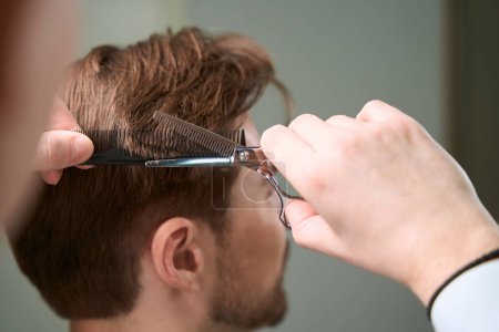 Téléchargez les photos : Vue recadrée sur les mains du coiffeur créant un nouveau look de cheveux du visiteur en utilisant des ciseaux et peigne dans la salle de salon - en image libre de droit