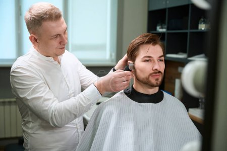Photo for Diligent hairdresser giving stylish haircut to his client by using electric clipper in modern salon - Royalty Free Image
