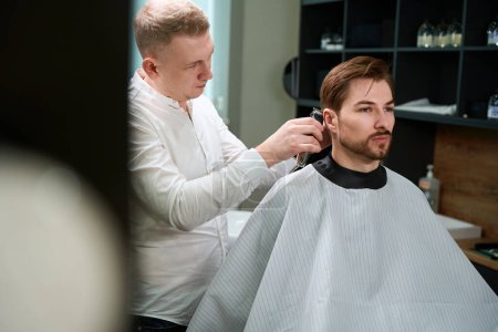 Photo for Handsome man in cutting hair cape sitting while professional hairdresser trimming him with barber machine indoors - Royalty Free Image