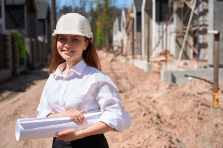 Photo for Smiling woman building engineer standing at construction site with blueprint at hands, working at building development, projecting houses - Royalty Free Image
