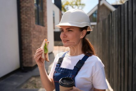 Téléchargez les photos : Ingénieur de bâtiment attrayant femme en casque et en général manger sandwich et boire du café à emporter pendant la pause déjeuner sur le chantier de construction, collation saine - en image libre de droit