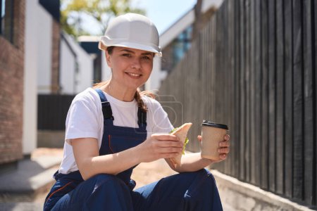 Téléchargez les photos : Femme souriante dans l'ensemble et hardhat a pause déjeuner, manger un sandwich savoureux et boire du café assis à l'extérieur sur le chantier de construction - en image libre de droit