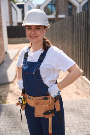 Téléchargez les photos : Ingénieure de la construction confiante en casque rigide et avec ceinture d'outils regardant la caméra et souriant, travaillant sur le chantier - en image libre de droit