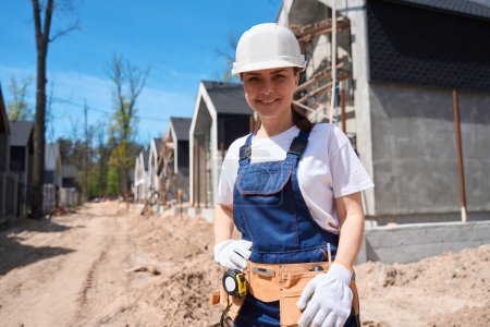 Téléchargez les photos : Femme souriante ingénieur du bâtiment en casque et gants de protection tenant la main sur la ceinture d'outils, prête à commencer les travaux sur le chantier - en image libre de droit