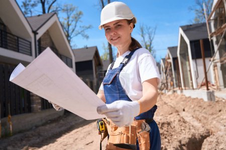 Photo for Forewoman in overall, hardhat and protective gloves arrived at construction site to studying blueprints and beginning work, paving slab laying - Royalty Free Image