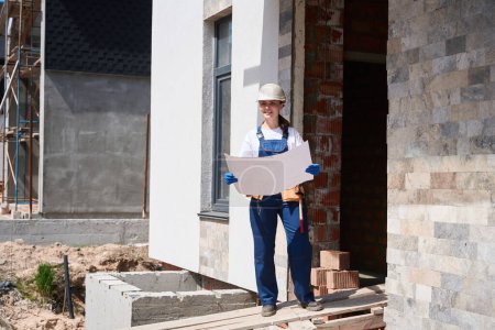 Téléchargez les photos : Entrepreneur femme positive debout près de la maison en construction entrée et regardant à travers le plan de construction, la planification des travaux de finition - en image libre de droit