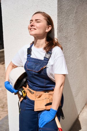 Téléchargez les photos : Portrait de femme ingénieure constructeur attirante dans l'ensemble et avec ceinture d'outils reposant et profitant de rayons de soleil chauds debout à l'extérieur, travaillant sur le chantier - en image libre de droit