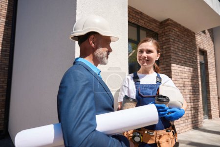 Téléchargez les photos : Architecte masculin sympathique parlant à une femme ingénieure attirante pendant la pause café, debout à l'extérieur près du bâtiment en construction - en image libre de droit