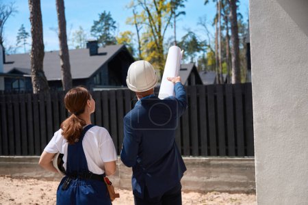 Photo for Back view male architect pointing at buildings, showing something to woman building engineer, colleagues discussing landscape design - Royalty Free Image