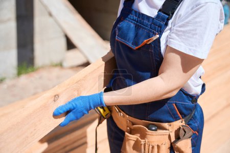 Téléchargez les photos : Gros plan femme constructeur en uniforme et ceinture à outils tenant poutre en bois, apportant du matériel préparé dans la maison en construction - en image libre de droit