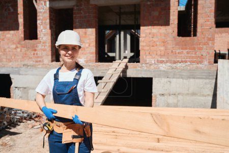 Foto de Mujer carpintero sosteniendo la viga larga, la elección de material adecuado para el suelo o el techo, la construcción de madera de construcción dentro de la casa de ladrillo, proyecto de diseño - Imagen libre de derechos