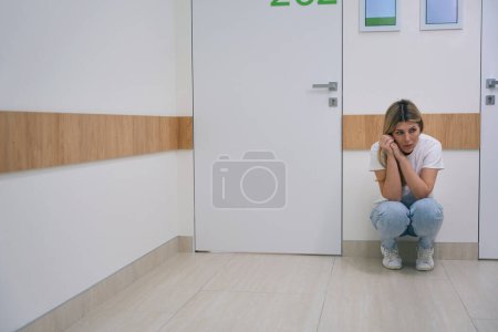 Téléchargez les photos : Femme blonde effrayée en jeans accroupi dans le couloir de l'hôpital, la femme est très bouleversée - en image libre de droit