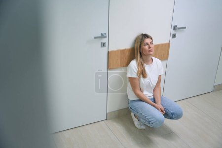 Foto de Joven mujer rubia en jeans y zapatillas de deporte está en cuclillas en el pasillo del hospital, la mujer está muy molesta - Imagen libre de derechos