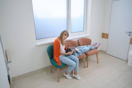 Téléchargez les photos : Fille avec sa mère installée dans la salle d'attente dans le couloir de l'hôpital, une femme console sa fille - en image libre de droit
