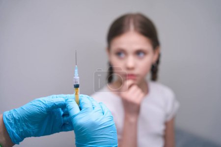 Photo for Medic in protective gloves holds a syringe with medicine in his hands, a young patient sits on a medical couch - Royalty Free Image