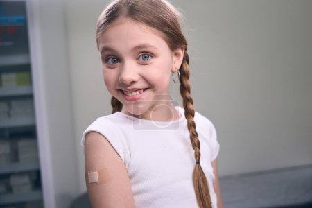Photo for Cheerful girl with pigtails and earrings in her ears sits in injection room, she has a band-aid on her arm - Royalty Free Image