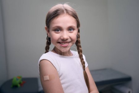 Photo for Cute girl with pigtails sits in an injection room, she has a band-aid on her arm - Royalty Free Image