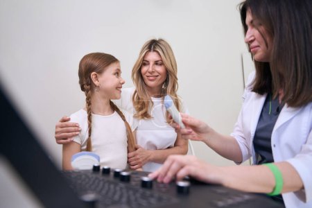 Photo for Doctor diagnostician communicates with a little patient in an ultrasound room, mother supports her daughter - Royalty Free Image