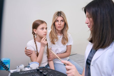 Téléchargez les photos : Femme inquiète avec sa fille lors d'un rendez-vous chez le médecin dans une clinique, le médecin introduit les patients à la machine à échographie - en image libre de droit