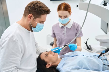 Téléchargez les photos : Dentiste professionnel en uniforme parlant à une cliente pendant le traitement des dents, tenant une perceuse dentaire, infirmière aidant tenant un éjecteur de salive, équipe de cliniques privées - en image libre de droit