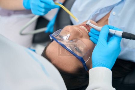 Téléchargez les photos : Stomatologue traitant les dents à la cliente dans des lunettes de protection avec une perceuse électrique qui tire de l'eau à la bouche, infirmière tenant un dispositif d'aspiration qui élimine la salive de la bouche - en image libre de droit