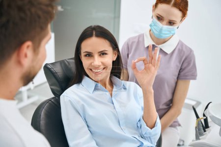 Téléchargez les photos : Belle femme souriante montrant signe de geste ok assis dans une chaise orthodontique après un traitement dentaire indolore et de qualité, dentiste qualifié et infirmière debout à proximité - en image libre de droit