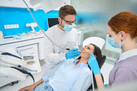 Téléchargez les photos : Équipe professionnelle de stomatologue traitant les dents des clientes, technicienne dentaire faisant un traitement de contrôle des racines, infirmière tenant un éjecteur de salive pendant que la patiente est allongée dans une chaise dentaire - en image libre de droit