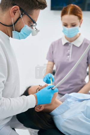 Photo for Focused doctor dental technician treating female client teeth, removing tooth decay before filling a cavity, nurse assisting holding suction device - Royalty Free Image