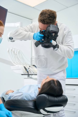 Photo for Dental technician making x-ray image of female client teeth and jaw holding in hands portable dental camera, exploring problems before treatment, private clinic service - Royalty Free Image