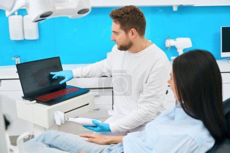 Téléchargez les photos : Technicien dentaire masculin pointant vers le moniteur numérique montrant des dents problématiques au client féminin après l'examen de la caméra interorale - en image libre de droit