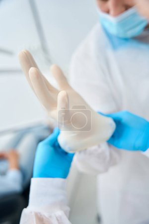 Téléchargez les photos : Infirmière aidant un technicien dentaire masculin hautement qualifié en uniforme à mettre des gants de stérille sur les mains avant le bilan de santé de la cavité buccale du client - en image libre de droit