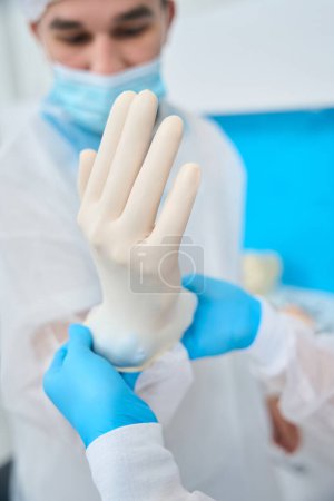Photo for Nurse assisting to male orthodontist in uniform and face mask, helping him to put on protective gloves, dentures or braces installation, aesthetic stomatology - Royalty Free Image