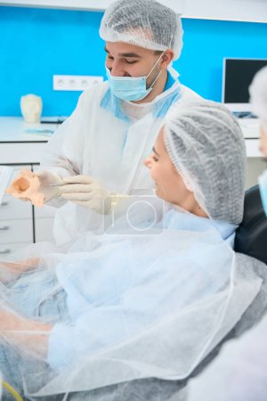 Foto de Hombre dentista se comunica con un paciente y le muestra un modelo de la mandíbula, un médico en uniforme médico - Imagen libre de derechos