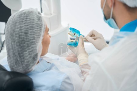 Téléchargez les photos : Patient examine le modèle avec des implants dentaires, le médecin utilise un masque de protection et des gants - en image libre de droit