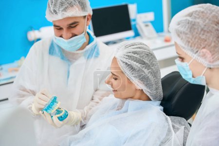 Téléchargez les photos : Femme examine une maquette avec des implants dentaires, un médecin et un assistant utilisent des masques de protection - en image libre de droit